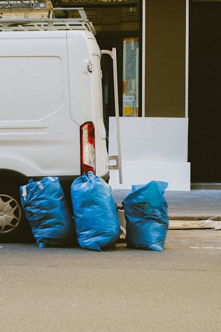 A large pile of mixed waste and rubbish bags is accumulated around a metal recycling bin labeled for paper and cardboard, situated on a paved area beside a row of parked cars in front of a commercial