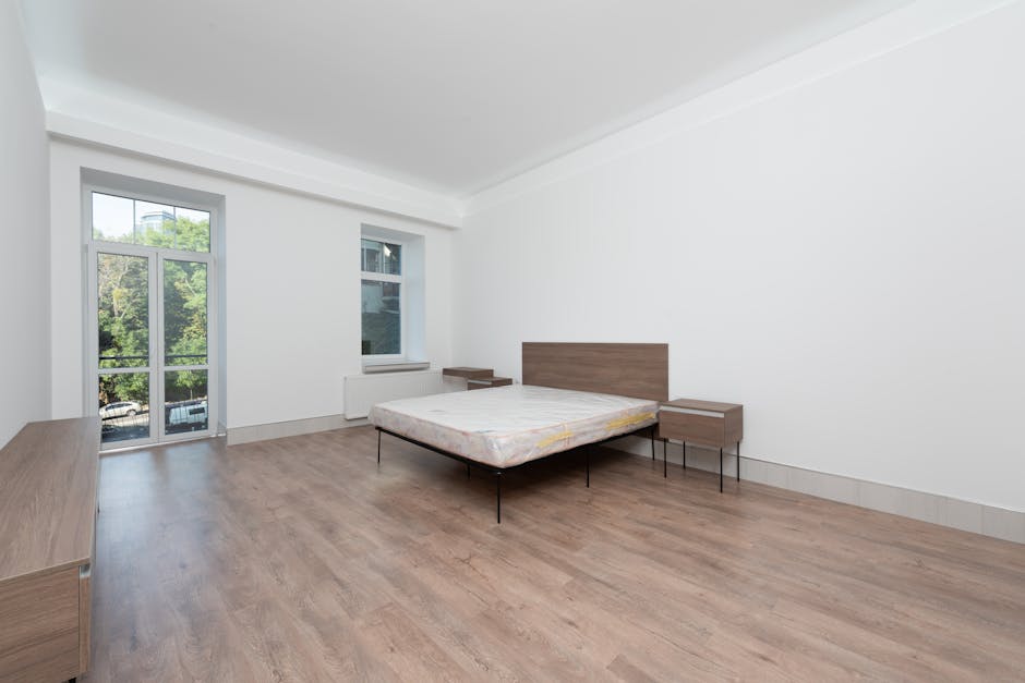 A minimalist bedroom scene featuring a double bed with a plain white mattress and a vertically tufted upholstered headboard in white leather or faux leather, positioned against a white wall. The room
