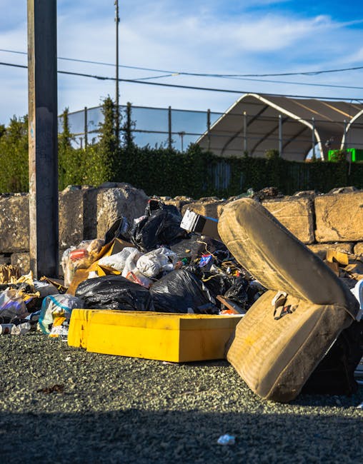 A cluttered outdoor scene showing a pile of discarded household items and debris in front of a damaged building. The pile includes white appliances such as a washing machine and possibly an oven, with